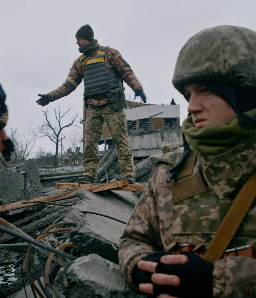 záběr z fronty s ozbrojeným vojákem v popředí a dvěma vojáky přenášejícími děti v pozadí